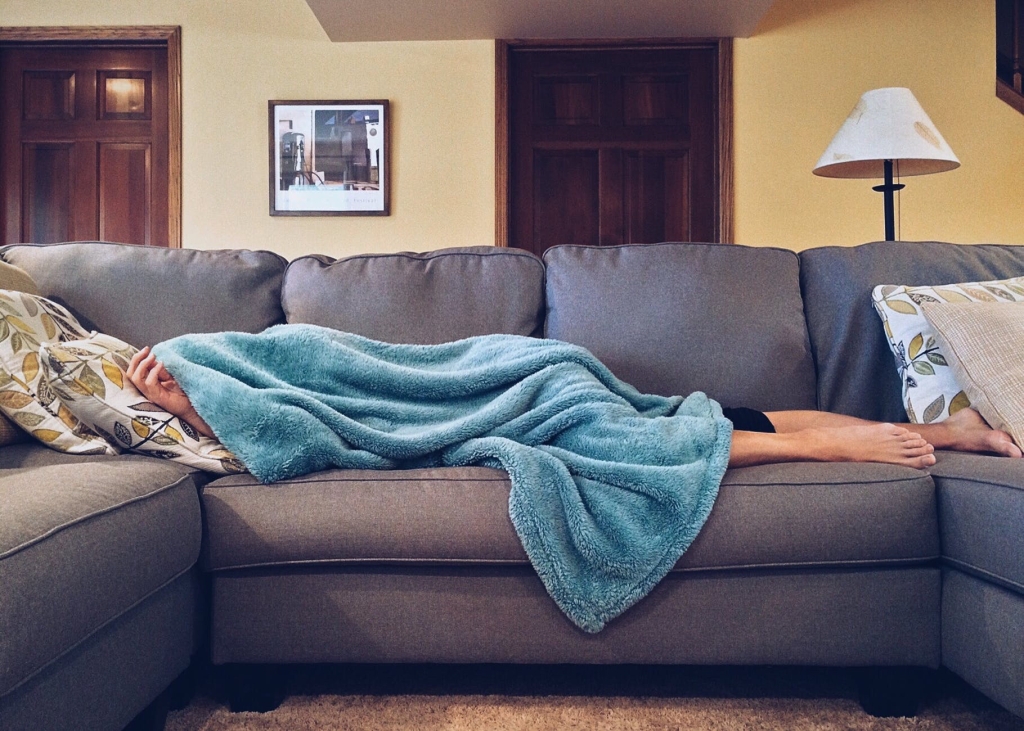 Photo of person lying down on a bluish-gray sofa with a blue blanket over their head