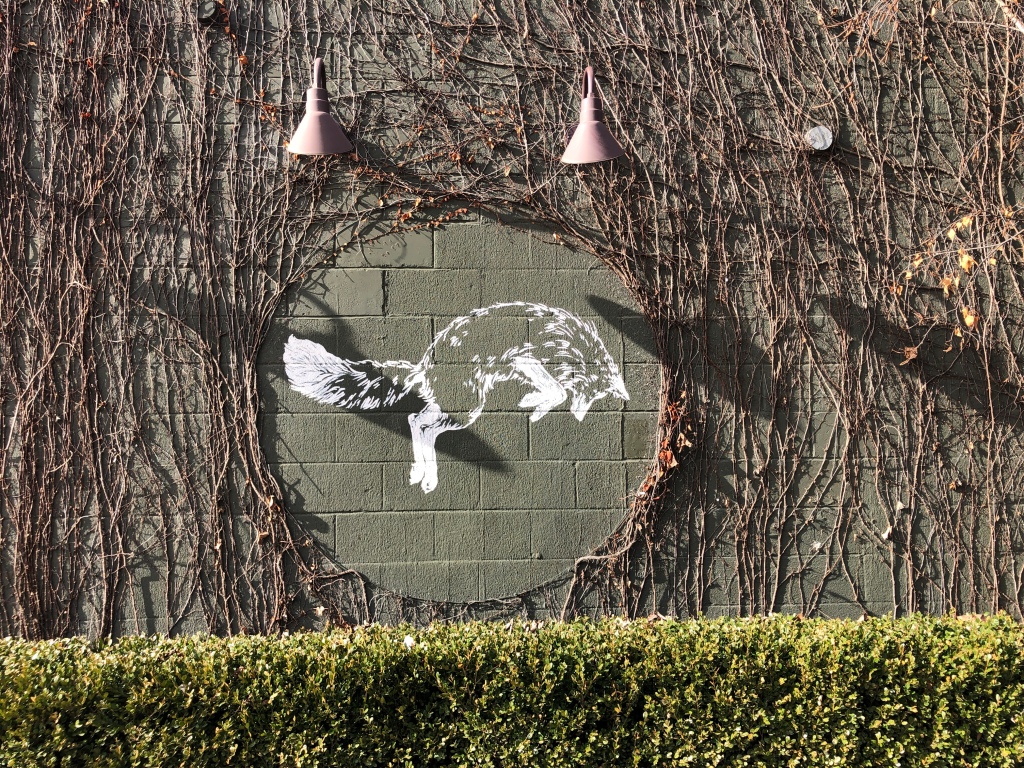 Photo of an illustration of a snow fox on a green brick wall with vines on the wall